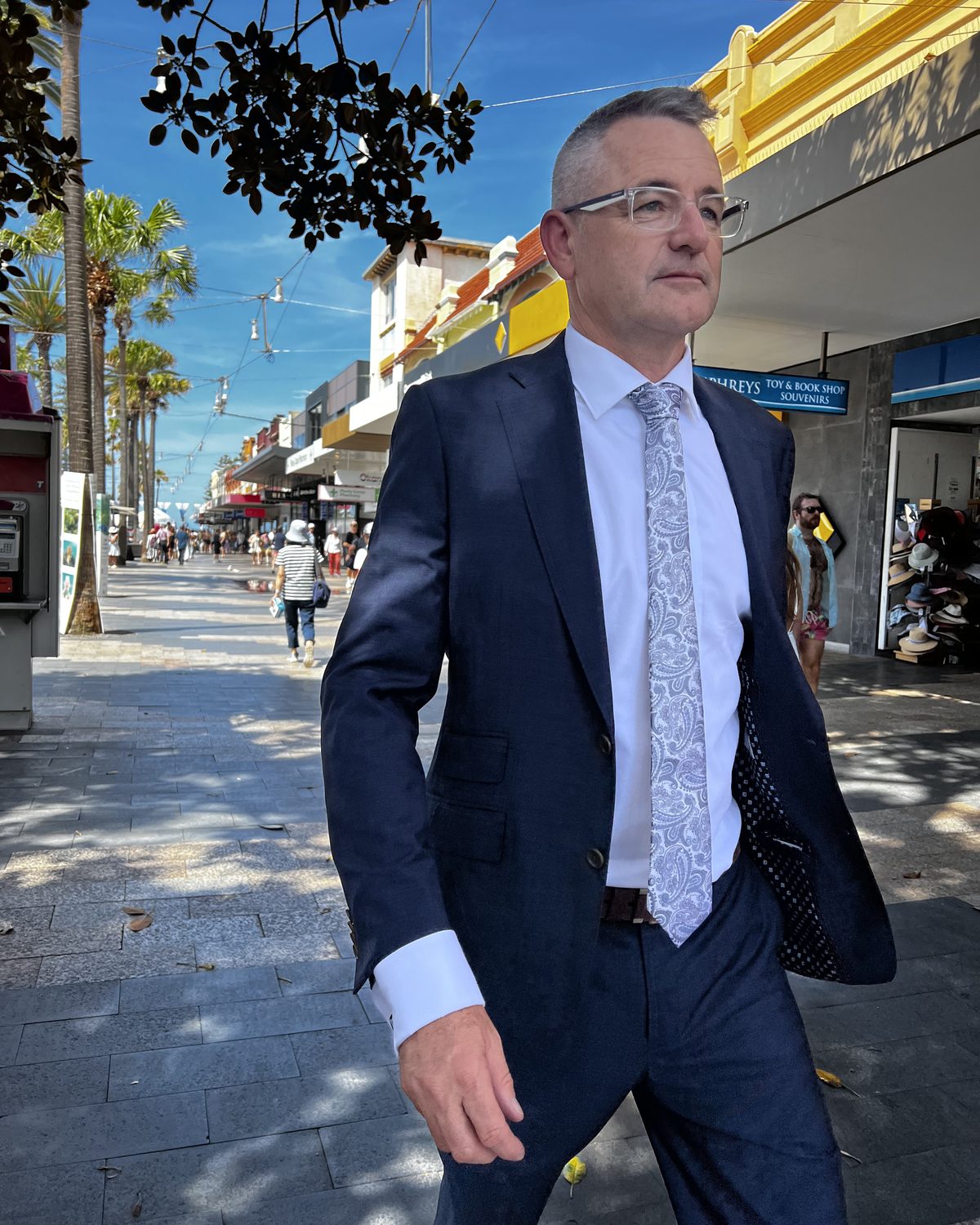 Andrew France walking along a sunny Sydney street in a navy suit
